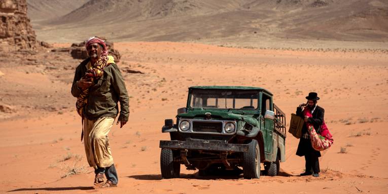 Ein Jeep steht in der Wüste. Ein Mann entfernt sich zu Fuß von ihm. Ein weiterer Mann packt noch seine Sachen.